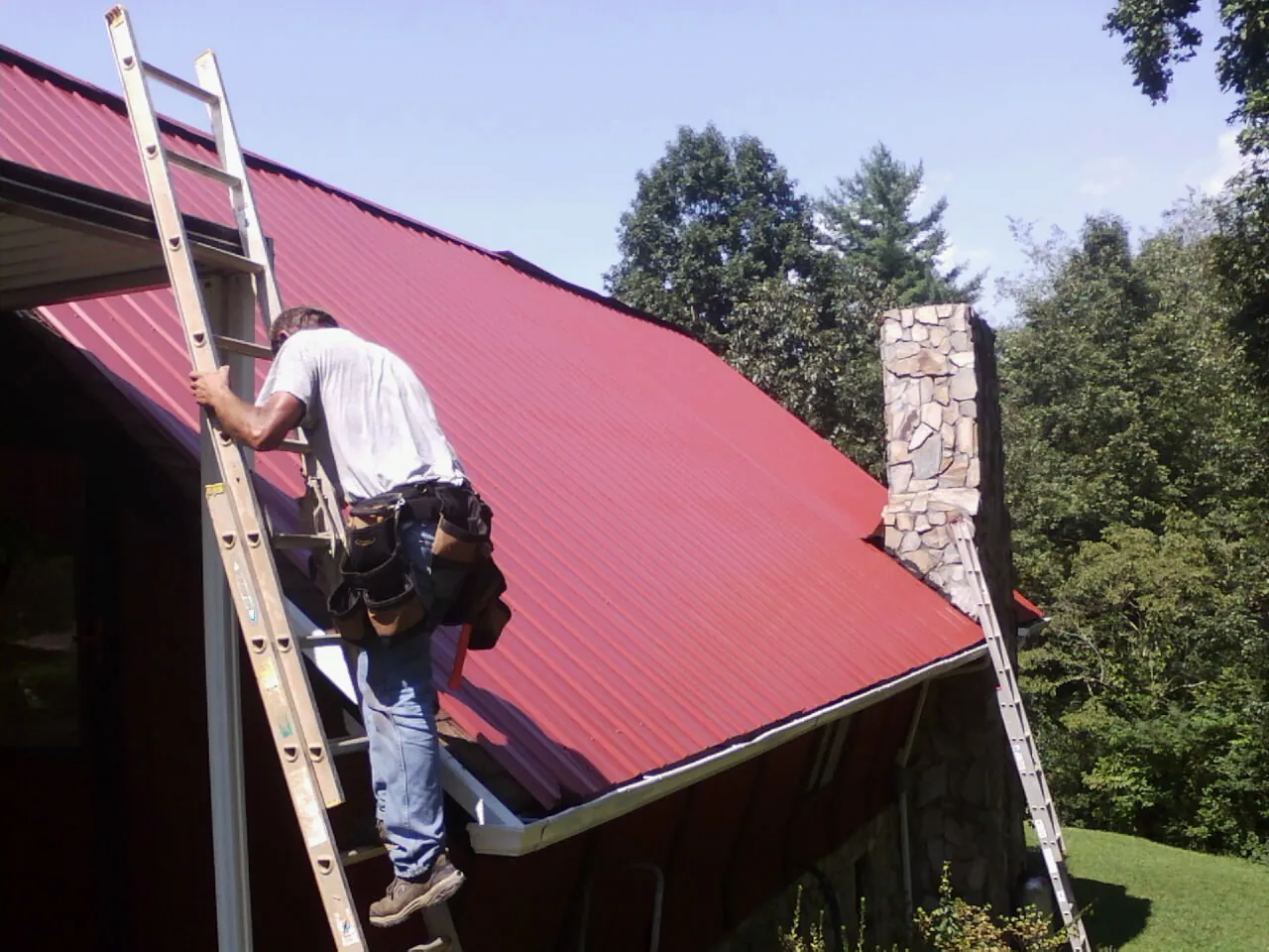 Roofer on a ladder beside a red metal roof and stone chimney — Soffit & Fascia Repair by Roofing Brothers in Lakeshore